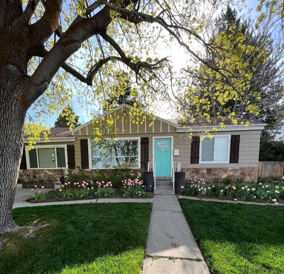 The exterior of a single story home that has a tan color on the main body of the house, white accented trim, and a teal front door.